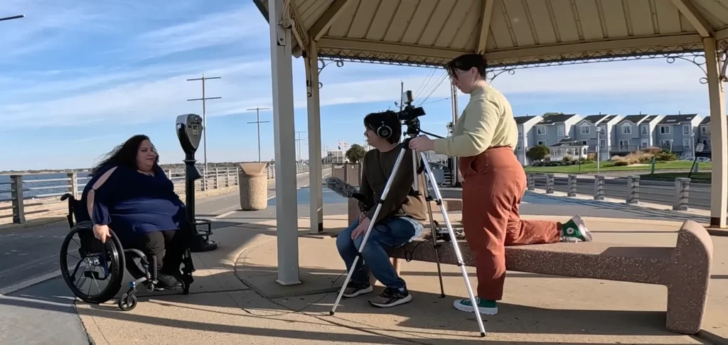A woman in a wheelchair sits in front of a man holding a microphone and a woman standing behind a camera atop a tripod. They are adjacent to a gazebo in a beachfront park.