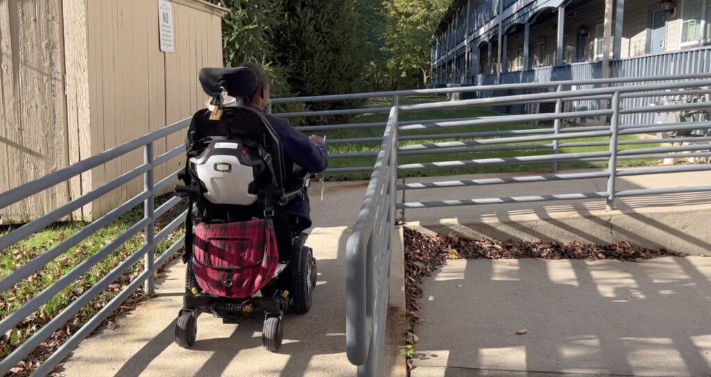 a dark skinned woman uses a power wheelchair while ascending a wooden ramp outside an apartment building