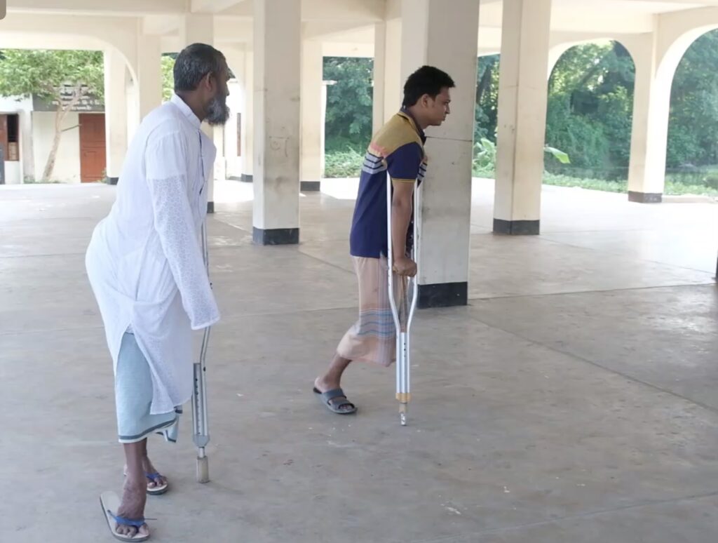 Two men stand in an open air pavilion. One steadies himself with a cane, the other, who does not have a left leg, balances himself with a pair of crutches.