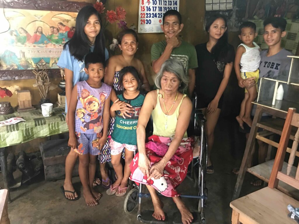 A Southeast Asian woman with grey hair sits in a wheelchair, surrounded by people of all ages