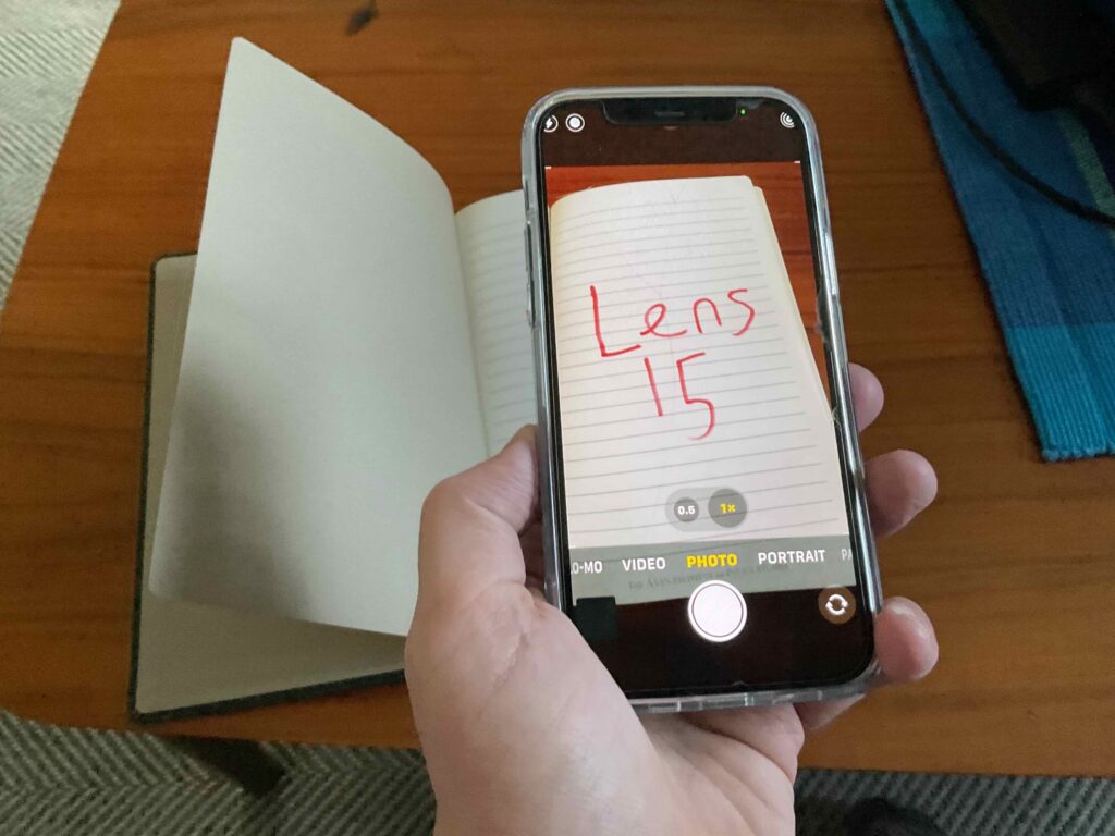 A smart phone is held over a notebook. On the screen, the words lens 15.